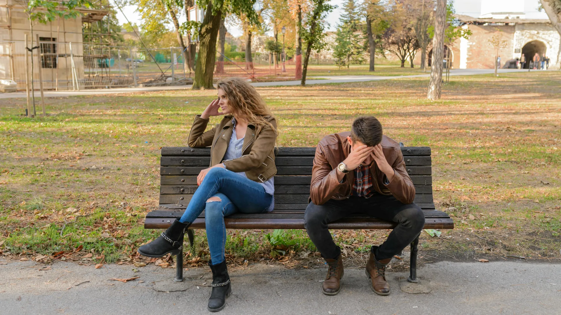 A couple are not interacting with other on a park bench. The female has her hand on her head turned away and the male has his hands in his head.